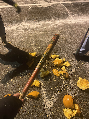 A photo of me and my friends smashing pumpkins on parking lot pavement. Both of my friends are out of frame, and in the foreground is me holding a baseball bat. Pumpkin remains lay on the ground.